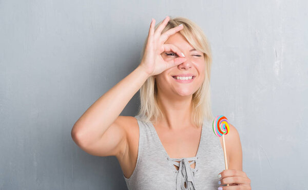 Caucasian adult woman over grey grunge wall eating candy lollipop with happy face smiling doing ok sign with hand on eye looking through fingers