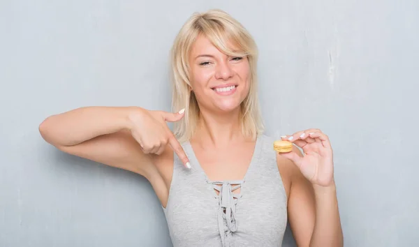 成人白种女人在垃圾灰色墙壁吃 Macaron 与惊奇面孔指向自己手指 — 图库照片