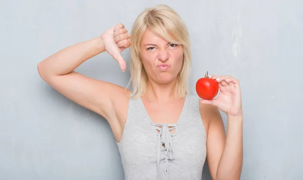 成年白种女人在灰墙上抱着新鲜的西红柿与愤怒的脸 消极的迹象显示不喜欢拇指下来 拒绝概念 — 图库照片