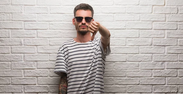 Genç yetişkin adam mutsuz ve kızgın ret gösterilen ve başparmak aşağı hareketi ile negatif seyir beyaz tuğla duvarın üzerinden duran güneş gözlüğü takıyor. Kötü ifade.