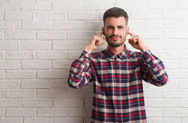 Yüksek sesle müzik gürültü rahatsız ifade parmaklarıyla kulaklarıyla kapsayan beyaz tuğla duvarın üzerinden ayakta genç yetişkin adam. Sağır kavramı.