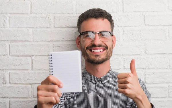 年轻成年人手持笔记本站在白色砖墙上快乐的笑着做 拇指与手指 优秀的标志 — 图库照片