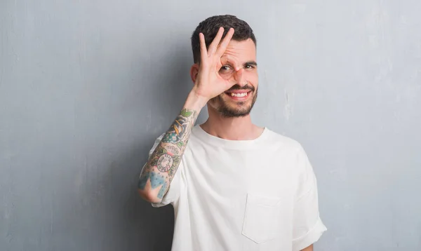 Young adult man standing over grey grunge wall with happy face smiling doing ok sign with hand on eye looking through fingers
