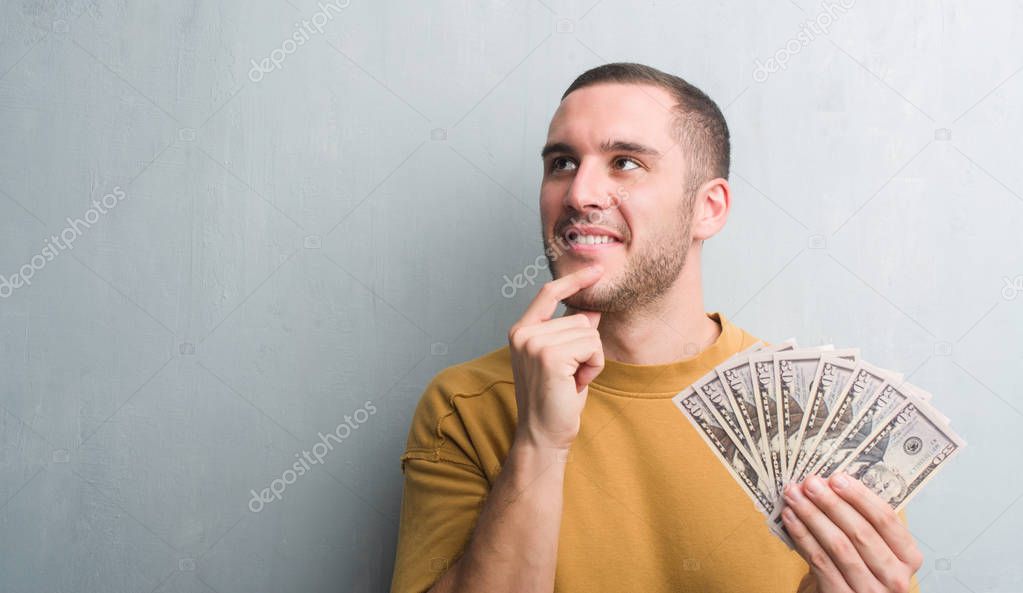 Young caucasian man over grey grunge wall holding dollars serious face thinking about question, very confused idea