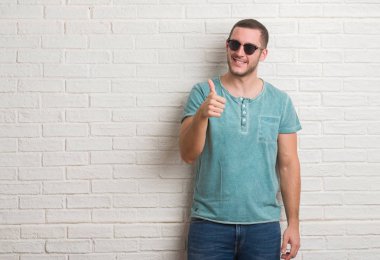 Ok işareti, başparmak ile parmak, mükemmel işareti yapıyor kocaman bir gülümseme ile mutlu güneş gözlüğü takmış beyaz tuğla duvarın üzerinden ayakta beyaz delikanlı