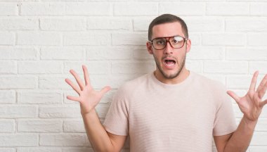 Genç beyaz adam ayakta üzerinde beyaz tuğla duvar büyük gülümseme ile çığlık zaferini kutluyor ve ellerini kaldırdı giyen gözlük çok mutlu ve heyecanlı, kazanan ifade