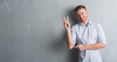 Genç redhead iş adamı mutlu yüz zafer işareti yaparken kameraya göz kırpıyor ile gülümseyen gri grunge duvarın üzerinden. İki numaralı.