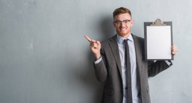 Genç redhead iş adamı Pano çok mutlu el ve parmak ile tarafına işaret tutan gri grunge duvarın üzerinden