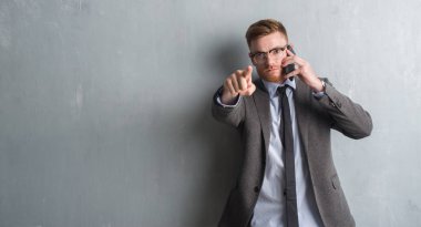 Genç redhead iş adamı kamera ve size parmak ile işaret telefonda konuşurken gri grunge duvarın üzerinden, el işareti, pozitif ve kendinden emin bir jest ön