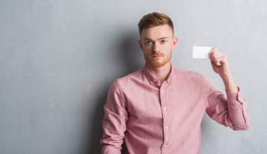 Genç Kızıl saçlı adam boş tutan gri grunge duvarın üzerinden ziyaret Kart kendine güvenen bir ifade ile acımak yüz ciddi düşünme