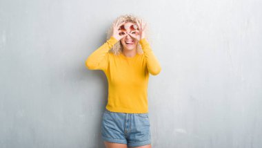 Sarışın genç kadın dilini, parmaklarının arasından baktığını yapışmasını dürbün gibi ok jest yapıyor grunge gri arka plan üzerinde Kıvırcık saçlı. Deli ifade.