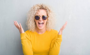 Young blonde woman with curly hair over grunge grey background celebrating mad and crazy for success with arms raised and closed eyes screaming excited. Winner concept clipart