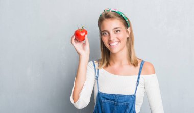 Güzel genç kadın ayakta ve diş gösterilen kendine güvenen bir gülümsemeyle gülümseyerek mutlu bir yüz ile taze domates yeme grunge gri duvarın üzerinden