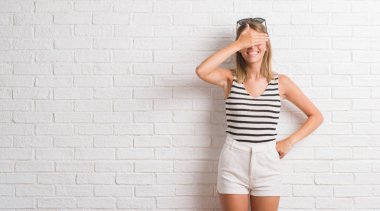 Genç güzel sarışın kadın gülümsüyor ve gözleri için sürpriz kapsayan yüz el ile gülüyor beyaz tuğla duvarın üzerinden. Kör kavramı.