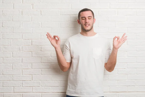 年轻的白种人站在白色砖墙放松和微笑闭上眼睛做冥想手势用手指 瑜伽理念 — 图库照片