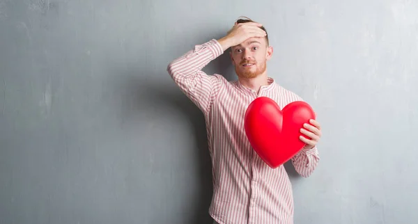 Young Redhead Man Grey Grunge Wall Holding Red Heart Stressed — Stock Photo, Image