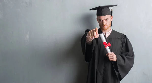 灰色のグランジの赤毛の若い男壁着て大学院制服カメラと指で指している学位を保持 手に署名 正面からポジティブで自信のジェスチャー — ストック写真