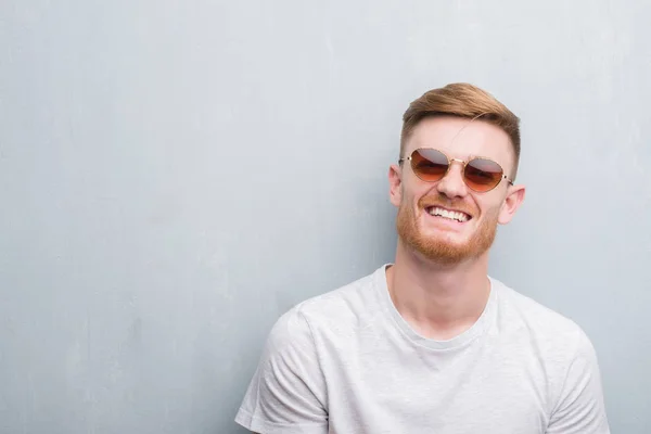 Young redhead man over grey grunge wall wearing retro sunglasses with a happy face standing and smiling with a confident smile showing teeth