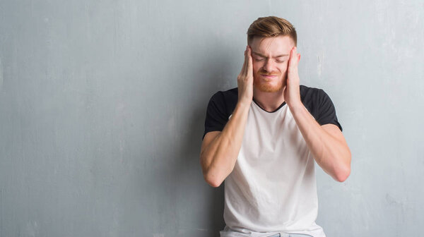 Young redhead man over grey grunge wall with hand on head for pain in head because stress. Suffering migraine.