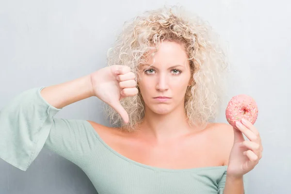 灰色背景的年轻金发女郎吃粉红色甜甜圈与愤怒的脸 消极的迹象显示不喜欢拇指向下 拒绝概念 — 图库照片