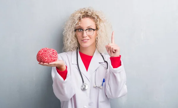 Jovem Mulher Médica Loira Sobre Grunge Cinza Parede Segurando Cérebro — Fotografia de Stock