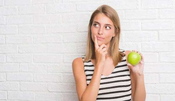 美丽的少妇在白色砖墙吃绿色苹果严肃的面孔思考问题 非常迷惑想法 — 图库照片
