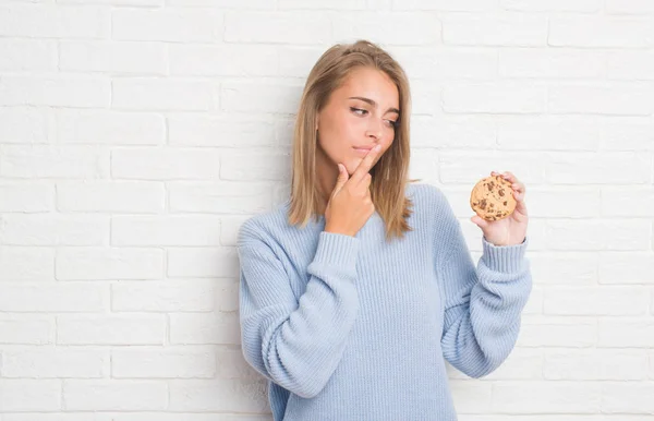 チョコレート チップ クッキー深刻な顔を考えて質問 非常に混乱して考えを食べる白いレンガの壁に美しい若い女性 — ストック写真