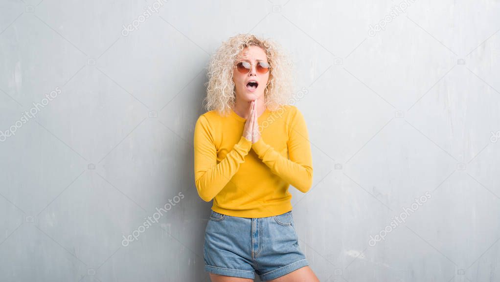 Young blonde woman with curly hair over grunge grey background begging and praying with hands together with hope expression on face very emotional and worried. Asking for forgiveness. Religion concept.