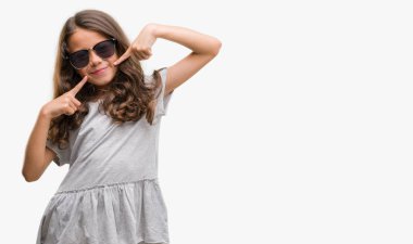 Esmer Latin kız kendinden emin gösterilen ve işaret parmakları diş ve ağız ile gülümseyen güneş gözlüğü takıyor. Sağlık kavramı.