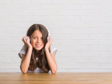 Ayakta ve diş gösterilen kendine güvenen bir gülümsemeyle gülümseyerek mutlu bir yüz ile kulaklıklar giyen masada oturan genç İspanyol çocuk