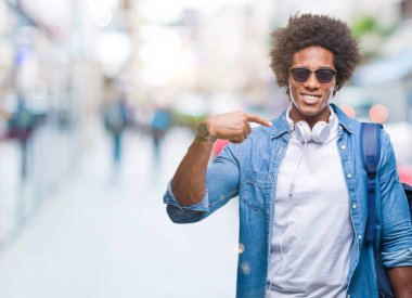 Afro Amerikalı adam kendisi için sürpriz yüz işaret parmağı ile izole arka plan üzerinde kulaklık ve sırt çantası giyiyor