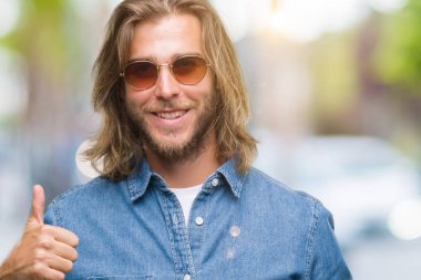 Genç yakışıklı adam uzun saçlı hareketi el ile mutlu yaşasın yapıyor izole arka plan üzerinde güneş gözlüğü takıyor. Kameraya bakarak başarı gösteren ile ifade onaylama.