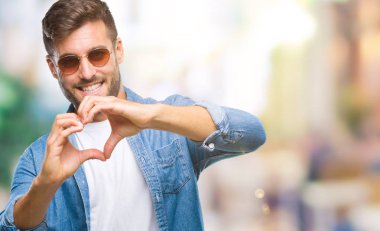 Genç yakışıklı adam aşık kalp simgesi ve elleriyle şekil gösteren gülümseyen izole arka plan üzerinde güneş gözlüğü takıyor. Romantik kavramı.