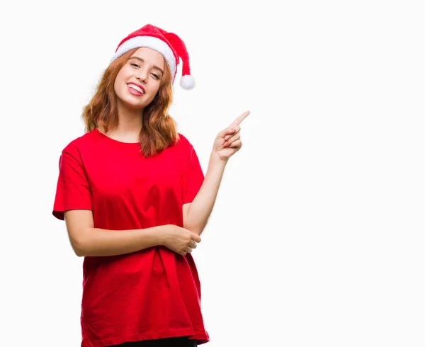 若くてきれいな女性の顔に大きな笑みを浮かべてクリスマス帽子をかぶっている孤立した背景にカメラを見ている側の手と指で指す — ストック写真
