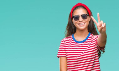 Genç güzel Arap kadın mutlu yüz zafer işareti yaparken kameraya göz kırpıyor ile gülümseyen izole arka plan üzerinde güneş gözlüğü takıyor. İki numaralı.