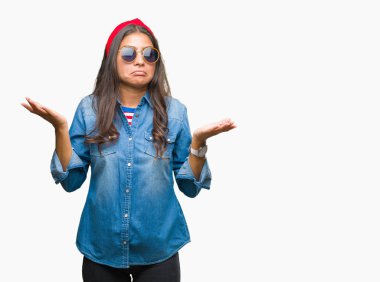 Genç güzel Arap kadın üzerinde güneş gözlüğü takıyor arka plan clueless ve şaşkın bir ifadeyle kollar ve eller kaldırdı izole. Şüphe kavramı.