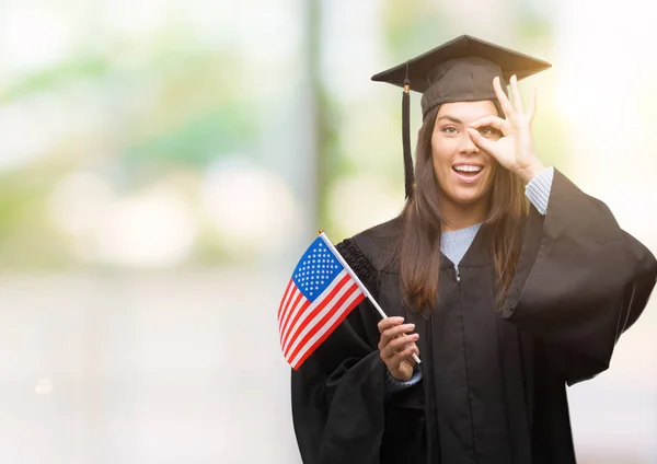 Young hispanic woman wearing graduated uniform holding flag of america with happy face smiling doing ok sign with hand on eye looking through fingers