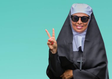 Orta yaş üst düzey Katolik Rahibe kadın mutlu yüz zafer işareti yaparken kameraya göz kırpıyor ile gülümseyen izole arka plan üzerinde güneş gözlüğü takıyor. İki numaralı.