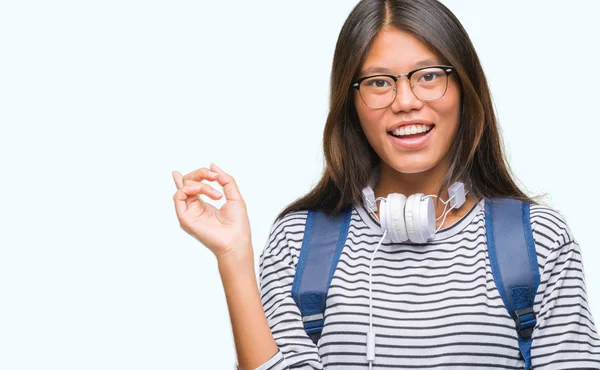 若いアジア学生の女性の顔に大きな笑みを浮かべて隔離された背景の上身に着けているヘッドフォンとバックパック カメラを見て側に手と指を指して — ストック写真