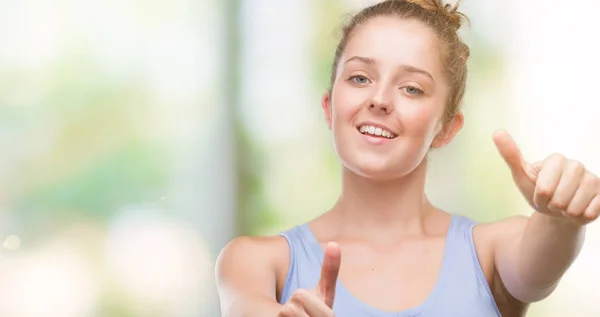 Young blonde woman approving doing positive gesture with hand, thumbs up smiling and happy for success. Looking at the camera, winner gesture.