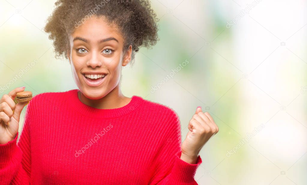 Young afro american woman eating chocolate macaron over isolated background screaming proud and celebrating victory and success very excited, cheering emotion