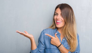 Grunge gri duvar hayrete ve el ile sunulması ve parmak ile işaret ederken kameraya gülümseyen denim kıyafeti giymiş genç yetişkin kadın.