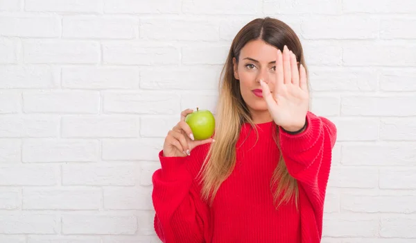 白色砖墙的年轻成年妇女吃新鲜绿色苹果用张开手做停止标志以严肃和自信的表达 防御姿态 — 图库照片