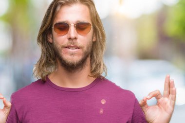 İzole arka plan üzerinde güneş gözlüğü takmış uzun saçlı yakışıklı delikanlı rahatla ve gülümseyen gözlerle kapalı parmak jestle meditasyon yapıyor. Yoga kavramı.