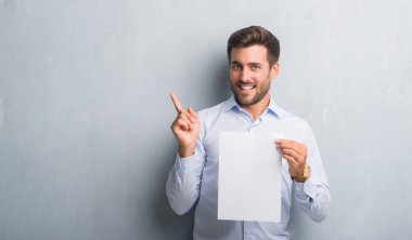 Yakışıklı genç gri grunge duvar holding boş kağıt sayfası üzerinden sözleşme çok mutlu el ve parmak ile işaret yan