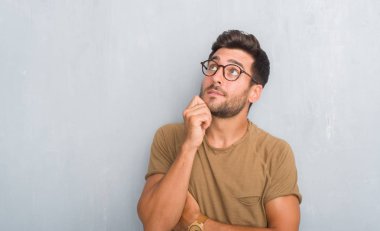 Yakışıklı genç adam gözlüklü el ile çene düşünme hakkında soru, dalgın ifade üzerine gri grunge duvarın üzerinden. Düşünceli yüzüyle gülümsüyor. Şüphe kavramı.