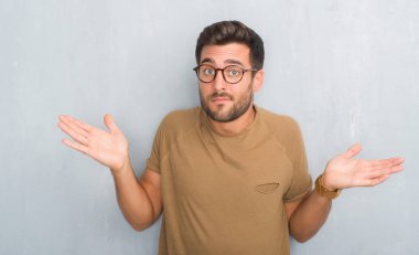 Yakışıklı genç gözlük clueless ve şaşkın bir ifadeyle kollar ve eller kaldırdı giyen gri grunge duvarın üzerinden. Şüphe kavramı.
