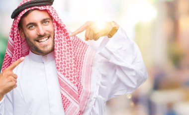 Keffiyeh kendine güvenen gösterilen ve işaret parmakları diş ve ağız ile gülümseyen izole arka plan üzerinde giyen genç yakışıklı adam. Sağlık kavramı.