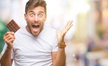 Genç yakışıklı adam büyük gülümseme ile çığlık zaferini kutluyor ve ellerini kaldırdı izole arka plan çok mutlu ve heyecanlı, kazanan ifade üzerinde çikolata yeme