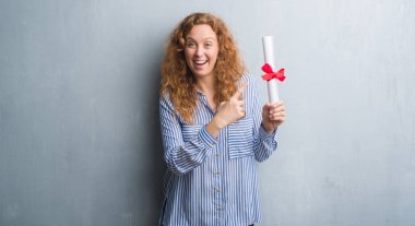 Genç redhead iş kadını diploması çok mutlu el ve parmak ile işaret tutan gri grunge duvarın üzerinden